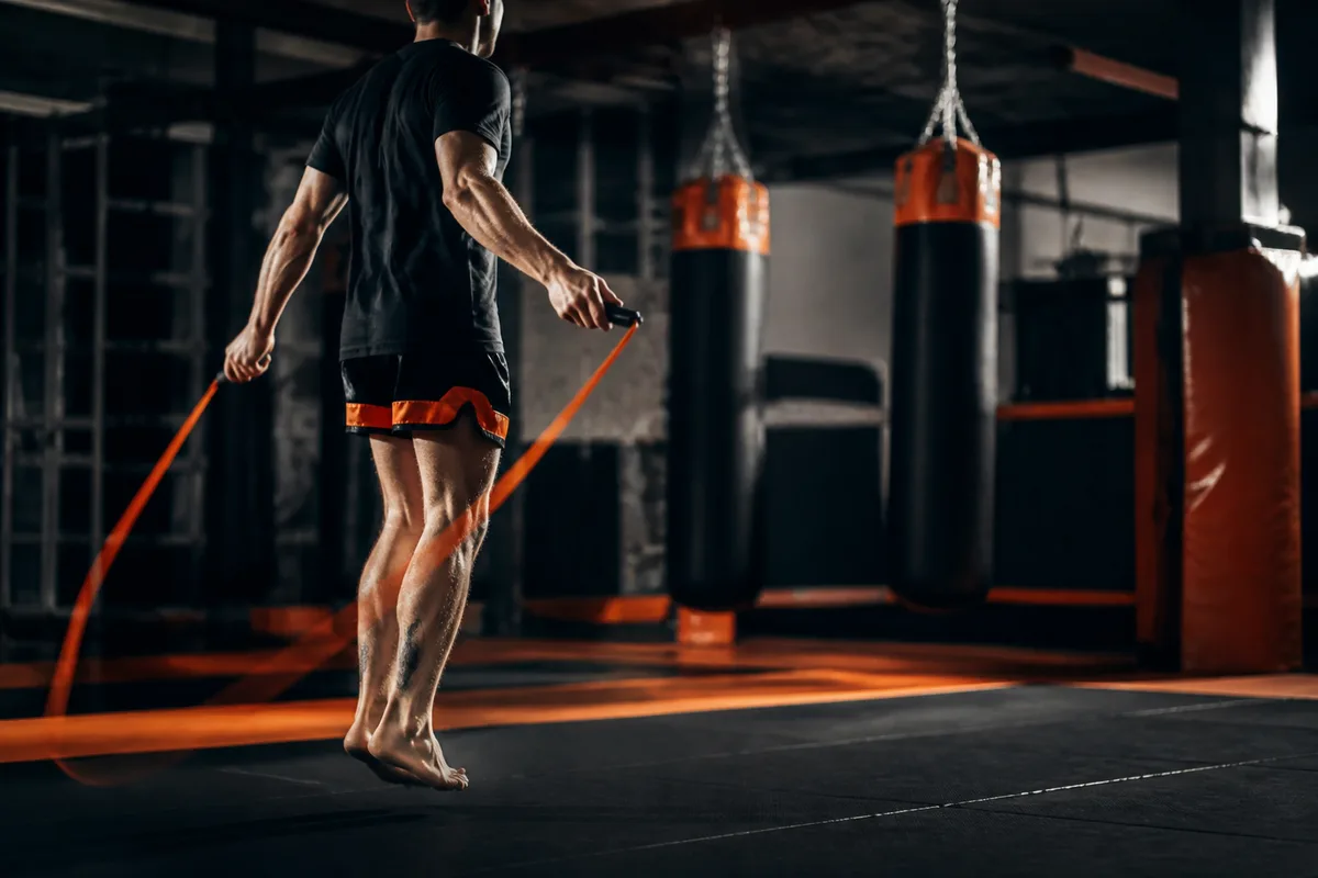 Un pratiquant débutant saute à la corde dans une salle d’entraînement, sacs de frappe suspendus en arrière-plan et lumière rasante