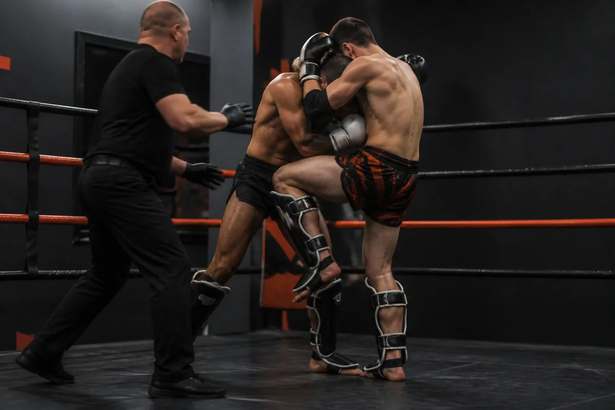Deux boxeurs de muay-thaï en position de clinch au centre d&rsquo;un ring, l&rsquo;arbitre à un pas d&rsquo;eux, prêt à séparer
